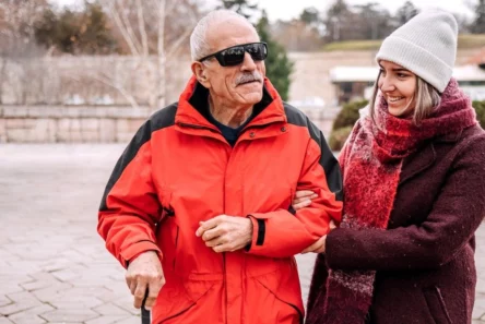 En ældre mand i rød jakke og solbriller går med en stok og smiler, mens en yngre kvinde i vinterfrakke og hue holder ham i armen og smiler til ham udendørs. Scenen viser, hvordan støtte til blinde – hvad enten det kommer fra omsorgsfulde ledsagere eller nye hjælpemidler som de AI-briller, kommunen stiller til rådighed – kan gøre hverdagens udflugter mere behagelige.