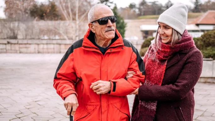 En ældre mand i rød jakke og solbriller går med en stok og smiler, mens en yngre kvinde i vinterfrakke og hue holder ham i armen og smiler til ham udendørs. Scenen viser, hvordan støtte til blinde – hvad enten det kommer fra omsorgsfulde ledsagere eller nye hjælpemidler som de AI-briller, kommunen stiller til rådighed – kan gøre hverdagens udflugter mere behagelige.