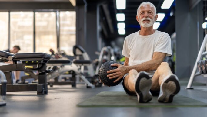 En ældre mand med hvidt skæg laver mavebøjninger, mens han holder en medicinbold i et fitnesscenter. Han har en hvid T-shirt, sorte shorts og sneakers på og smiler, mens han viser fordelene ved selv en 3 minutters træning. I baggrunden ses fitnessudstyr.