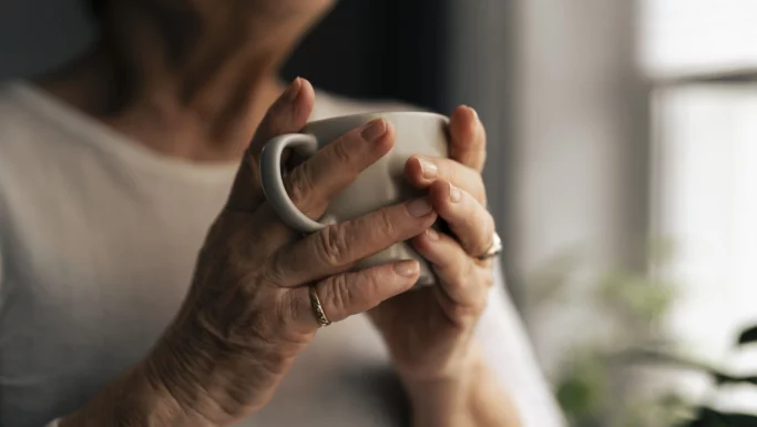 En person iført en lys skjorte holder en hvid kop i begge hænder tæt ved et vindue og nyder en varm drik, mens blødt naturligt lys belyser vedkommendes hænder og en del af ansigtet.