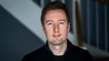 En mand med kort brunt hår og blå øjne, iført en sort trøje med lynlås, ser direkte ind i kameraet med et neutralt udtryk. Den slørede baggrund er præget af diagonale linjer og dæmpede farver, hvilket skaber en rolig stemning, der minder om en plan, der er ved at tage form – måske en, som Ældreministeren har drøftet.
