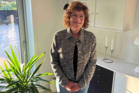 En kvinde med krøllet hår og briller smiler, mens hun står i et solfyldt rum tæt på en plante og fejrer sit 25-års jubilæum som plejemor, med hvide skabe, stearinlys og en lampe bag sig.