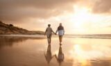 To mennesker, der holder hinanden i hånden, går barfodet langs en sandstrand ved solnedgang, tænker over deres fremtid og taler om pension. Himlen er delvis overskyet, og deres skygger kan ses på det våde sand.