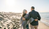 Et ældre par går arm i arm langs en sandstrand og ser på hinanden, mens solen skinner og palmerne står i baggrunden – de nyder deres gyldne år i ro og fred takket være gennemtænkte pensionsråd og en sikker aldersopsparing.