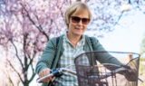 En smilende person iført solbriller og en ternet skjorte cykler på en cykel med en cykelkurv, mens der i baggrunden står blomstrende lyserøde træer.