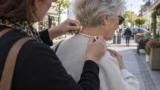 En kvinde hjælper en 78-årig ældre kvinde med at tage en perlekæde på, mens de står udendørs på en solrig gade i byen, der er fyldt med butikker, blomster og udstillinger af smukke guldhalskæder.