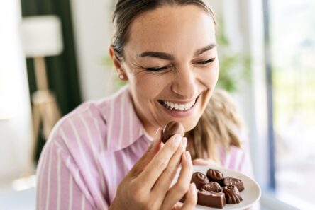 En smilende kvinde i en lyserød stribet bluse holder en tallerken med forskellige slags chokolader og er ved at spise en af dem, mens hun nyder godbidden og giver efter for sin sukkertrang i et lyst og hyggeligt rum.
