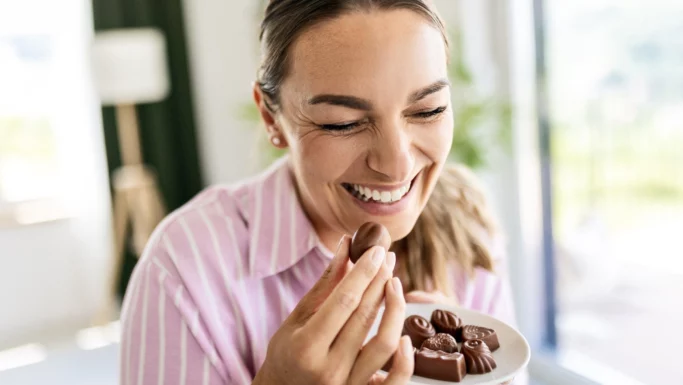 En smilende kvinde i en lyserød stribet bluse holder en tallerken med forskellige slags chokolader og er ved at spise en af dem, mens hun nyder godbidden og giver efter for sin sukkertrang i et lyst og hyggeligt rum.