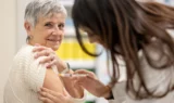 En ældre kvinde i en hvid trøje smiler, mens hun får en stærkere influenzavaccine indsprøjtet i overarmen af en sundhedsmedarbejder med langt brunt hår.