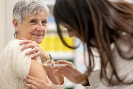 En ældre kvinde i en hvid trøje smiler, mens hun får en stærkere influenzavaccine indsprøjtet i overarmen af en sundhedsmedarbejder med langt brunt hår.