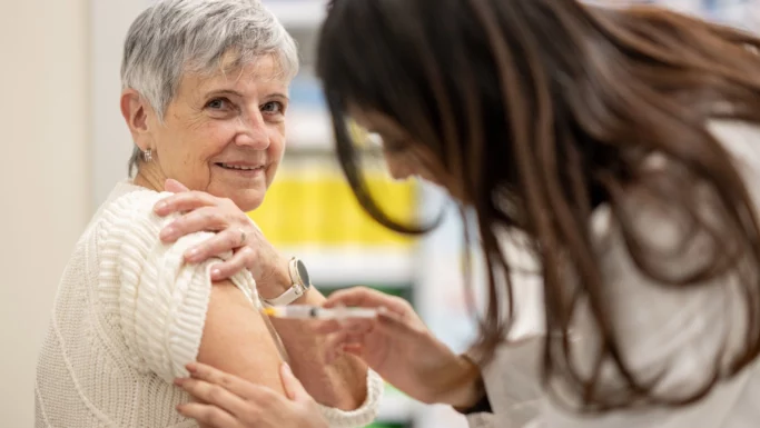 En ældre kvinde i en hvid trøje smiler, mens hun får en stærkere influenzavaccine indsprøjtet i overarmen af en sundhedsmedarbejder med langt brunt hår.