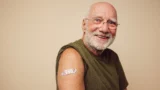 En smilende ældre mand med briller har en olivengrøn skjorte på og et farvestrålende plaster på overarmen, hvilket tyder på, at han netop er blevet vaccineret efter at have modtaget en invitation. En enkel beige baggrund.