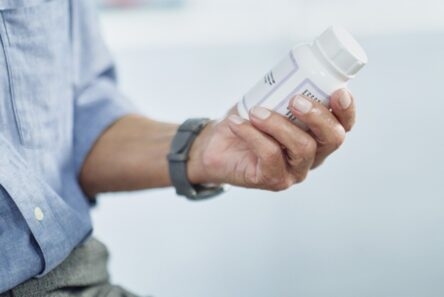 En person iført en blå skjorte og et gråt ur holder en hvid pilleæske i hånden og studerer etiketten nøje, måske for at tjekke oplysninger om medicin i forbindelse med åreforkalkning.