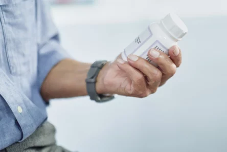 En person iført en blå skjorte og et gråt ur holder en hvid pilleæske i hånden og studerer etiketten nøje, måske for at tjekke oplysninger om medicin i forbindelse med åreforkalkning.