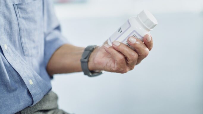 En person iført en blå skjorte og et gråt ur holder en hvid pilleæske i hånden og studerer etiketten nøje, måske for at tjekke oplysninger om medicin i forbindelse med åreforkalkning.