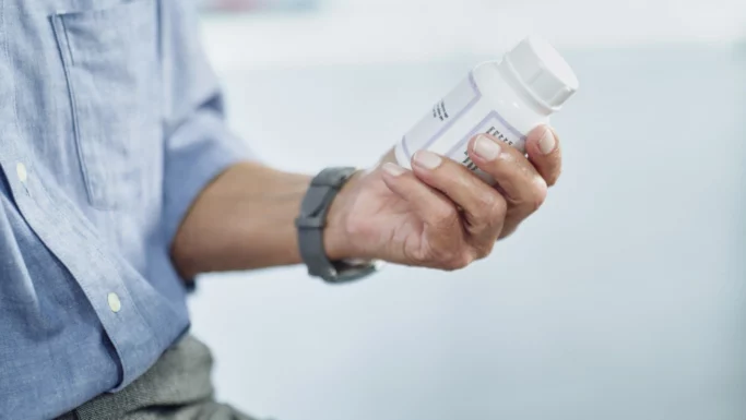 En person iført en blå skjorte og et gråt ur holder en hvid pilleæske i hånden og studerer etiketten nøje, måske for at tjekke oplysninger om medicin i forbindelse med åreforkalkning.