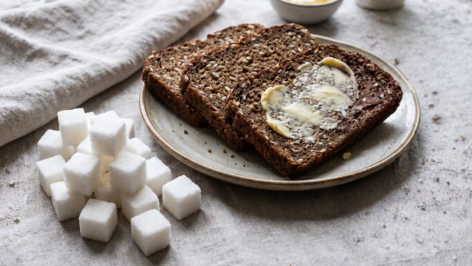 Tre skiver fuldkornsbrød på en tallerken, hvoraf den ene er smurt med smør, ved siden af en bunke sukkerterninger, der illustrerer kostens indvirkning på blodsukkeret.