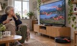 En ældre kvinde sidder i en stol med en fjernbetjening i hånden og ser fjernsyn med rødglødende AI-orme-grafik, der ligner lyset fra en robotstøvsuger, hvilket illustrerer bekymringerne omkring IT-sikkerhed.
