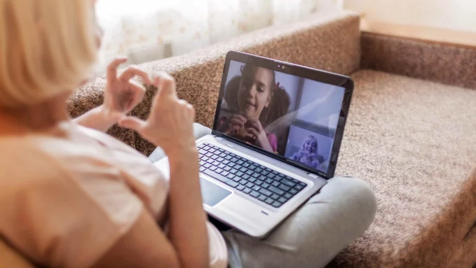 En person sidder på en trappe med en bærbar computer og videochatter med to børn. Et af børnene på skærmen efterligner håndbevægelser tæt på ansigtet, hvilket tyder på tegnsprog eller legende kommunikation. Scenen er oplyst af blødt indendørs lys.