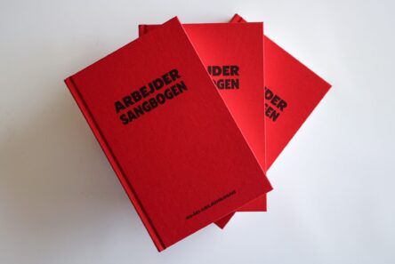 Tre røde indbundne eksemplarer af Arbejdersangbogen stablet på en hvid overflade.