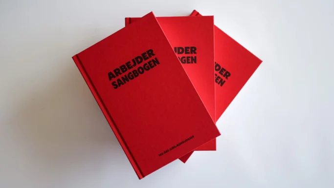 Tre røde indbundne eksemplarer af Arbejdersangbogen stablet på en hvid overflade.