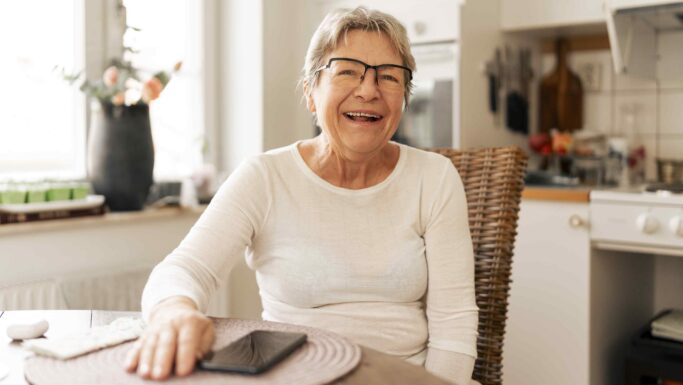 En smilende ældre kvinde med briller, der sidder ved et køkkenbord og tjekker sin ATP-pension på sin smartphone.