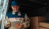 En erfaren leveringschauffør iført en blå kasket og uniform bruger en tablet, mens han står bag i en varevogn fyldt med papkasser.