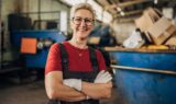 En kvinde med kort blondt hår og briller, iført en rød bluse og arbejdsoveralls, står med armene over kors i et lager og overvejer de økonomiske fordele ved at arbejde længere inden pensionering.