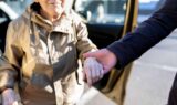 En person yder kørehjælp ved at holde en ældre kvinde i hånden, mens hun stiger ud af bilen, og dermed støtter hendes frihed.