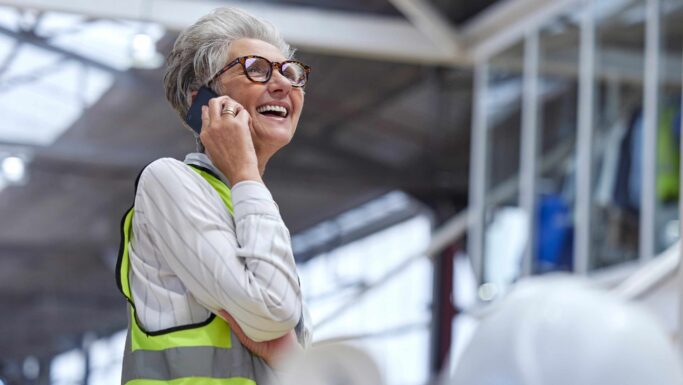 En smilende kvinde i en sikkerhedsvest taler i mobiltelefon, mens hun diskuterer nye regler om arbejde.