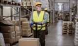 En lagermedarbejder iført en gul sikkerhedsvest og en sikkerhedshjelm betjener en pallevogn i en gang fyldt med papkasser, hvilket afspejler den indvirkning, som diskussionerne om bundskat og FH-afgiften har på branchen.
