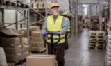 En lagermedarbejder iført en gul sikkerhedsvest og en sikkerhedshjelm betjener en pallevogn i en gang fyldt med papkasser, hvilket afspejler den indvirkning, som diskussionerne om bundskat og FH-afgiften har på branchen.