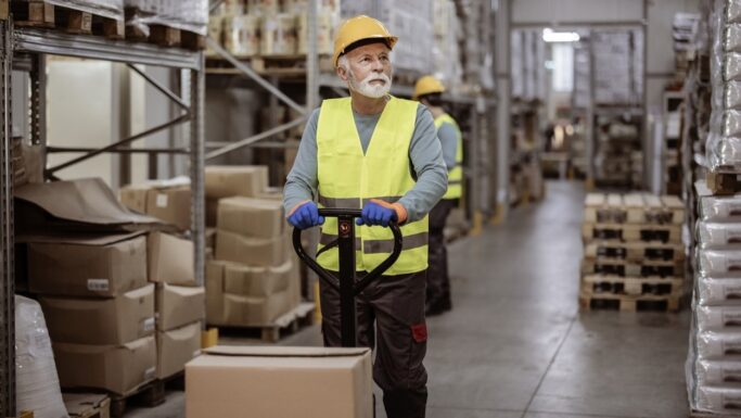 En lagermedarbejder iført en gul sikkerhedsvest og en sikkerhedshjelm betjener en pallevogn i en gang fyldt med papkasser, hvilket afspejler den indvirkning, som diskussionerne om bundskat og FH-afgiften har på branchen.