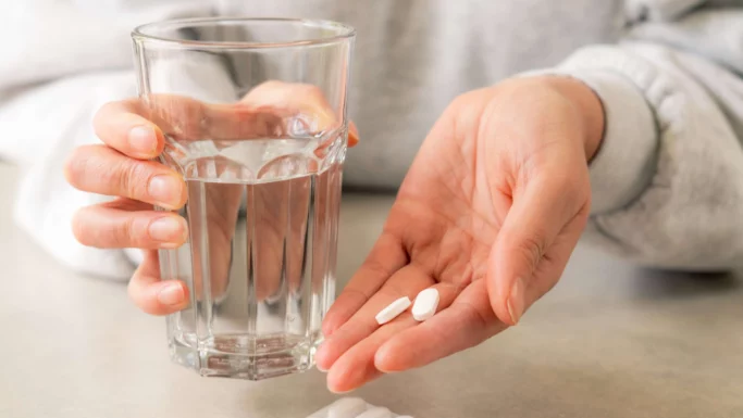 En person holder et glas vand i den ene hånd og to hvide paracetamol-piller i den anden for at lindre smerter.