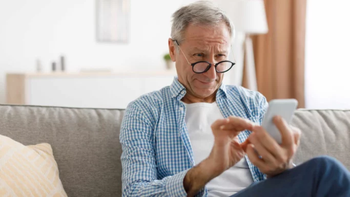 En ældre mand med briller, der kigger nøje på en MobilePay-skærm på sin smartphone, mens han sidder i en sofa.