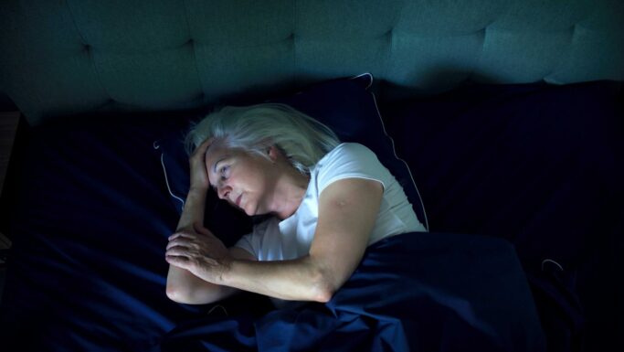 En ældre kvinde med gråt hår, der ligger vågen i sengen om natten og ser bekymret ud med hånden på panden på grund af søvnbesvær.