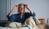 En smilende kvinde sidder i sengen og trækker sin søvnmaske op over panden, klar til en god nats søvn.
