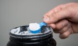 En hånd holder en blå plastikskovl fyldt med hvidt pulver over en åben sort beholder med kosttilskud.