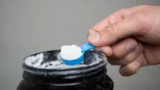 En hånd holder en blå plastikskovl fyldt med hvidt pulver over en åben sort beholder med kosttilskud.