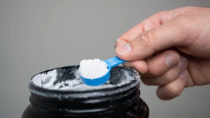 En hånd holder en blå plastikskovl fyldt med hvidt pulver over en åben sort beholder med kosttilskud.