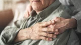 En person lægger en trøstende hånd på skulderen af en ældre mand, der lider af demens.