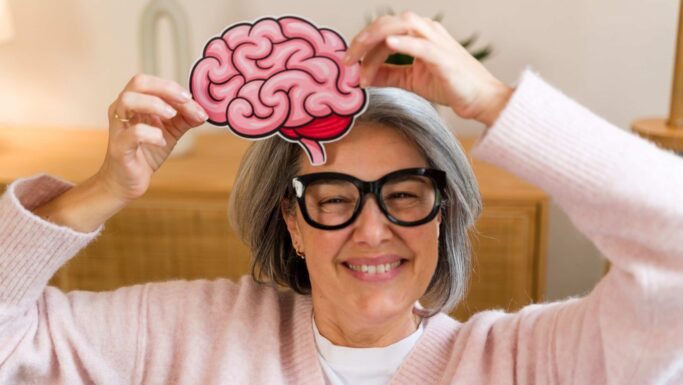 En kvinde med gråt hår og briller, der smiler, mens hun holder en tegneserieagtig udskæring af en hjerne over hovedet for at symbolisere hjerneforskning.