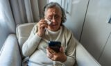 En ældre kvinde sidder i en lænestol og græder, mens hun kigger på en smartphone, hvor der vises en dødsannonce.