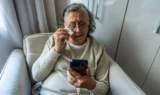 En ældre kvinde sidder i en lænestol og græder, mens hun kigger på en smartphone, hvor der vises en dødsannonce.