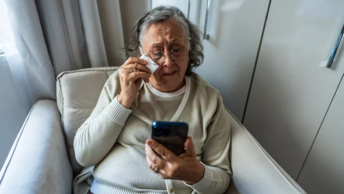 En ældre kvinde sidder i en lænestol og græder, mens hun kigger på en smartphone, hvor der vises en dødsannonce.
