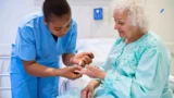 En sygeplejerske i blå kittel udfører en blodsukkermåling ved fingerprik på en ældre patient, der lider af en kronisk sygdom.