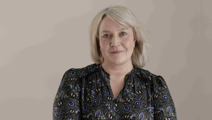 En middelaldrende præst med blondt hår, iført en grøn bluse med blomstermønster, ser mod kameraet.