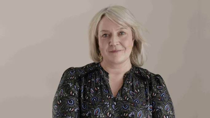 En middelaldrende præst med blondt hår, iført en grøn bluse med blomstermønster, ser mod kameraet.