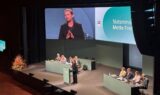 Mette Frederiksen taler fra et talerstol foran et publikum, mens et stort skærm viser et nærbillede af hende, da hun holder tale på en jobkonference for seniorer.