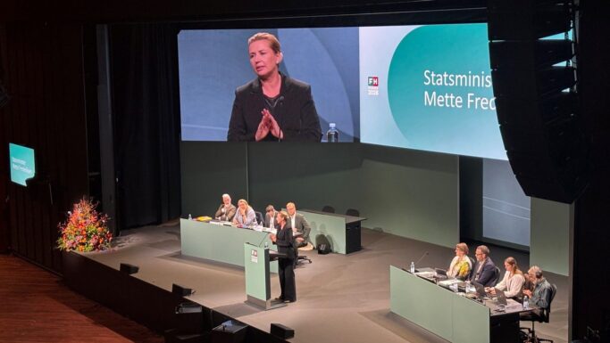 Mette Frederiksen taler fra et talerstol foran et publikum, mens et stort skærm viser et nærbillede af hende, da hun holder tale på en jobkonference for seniorer.